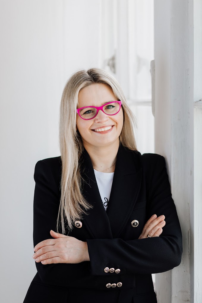 Portret uśmiechniętej kobiety w okularach i czarnej marynarce.